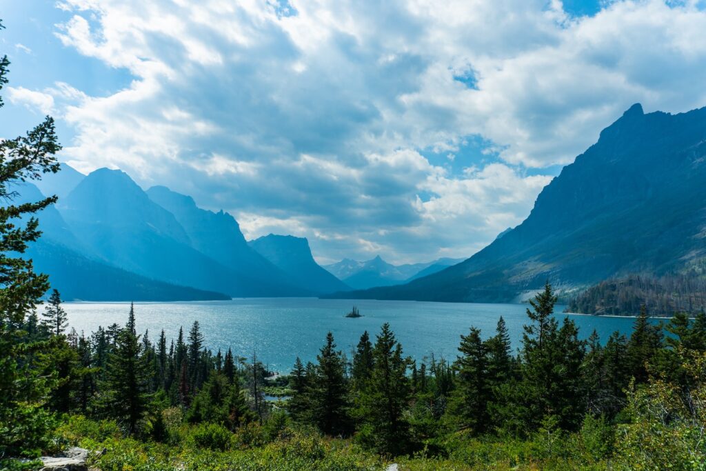 view of a lake surrounded by trees with a cloudy blue sky and mountains silhouetted in background at Glacier National Park in Montana