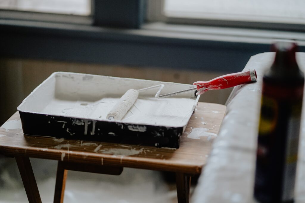 Paint roller covered in white paint sitting in a paint tray on top of a wooden table